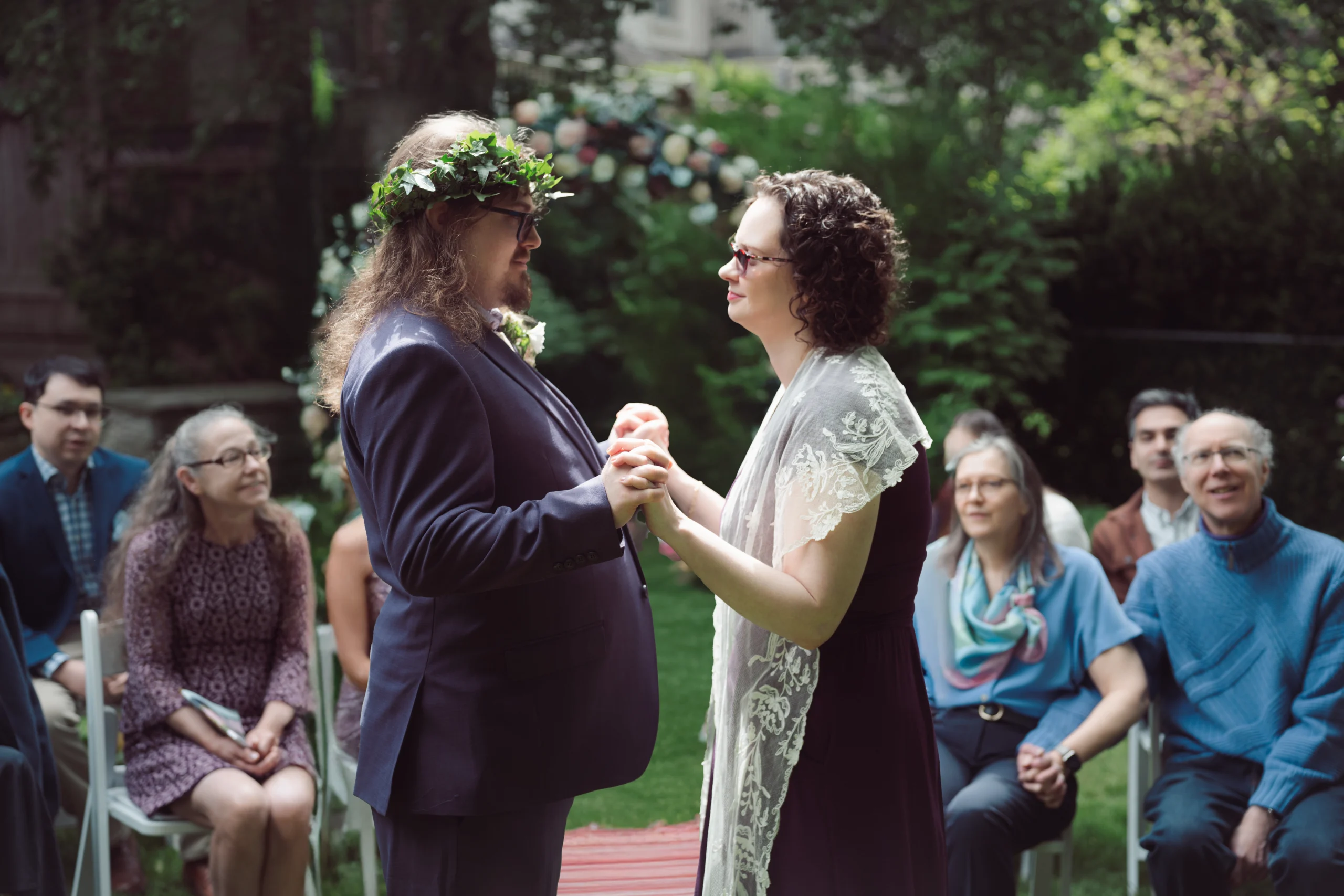 Bride and groom holding hands during ceremony during mid day while guests watch them in backyard full of grass and trees
