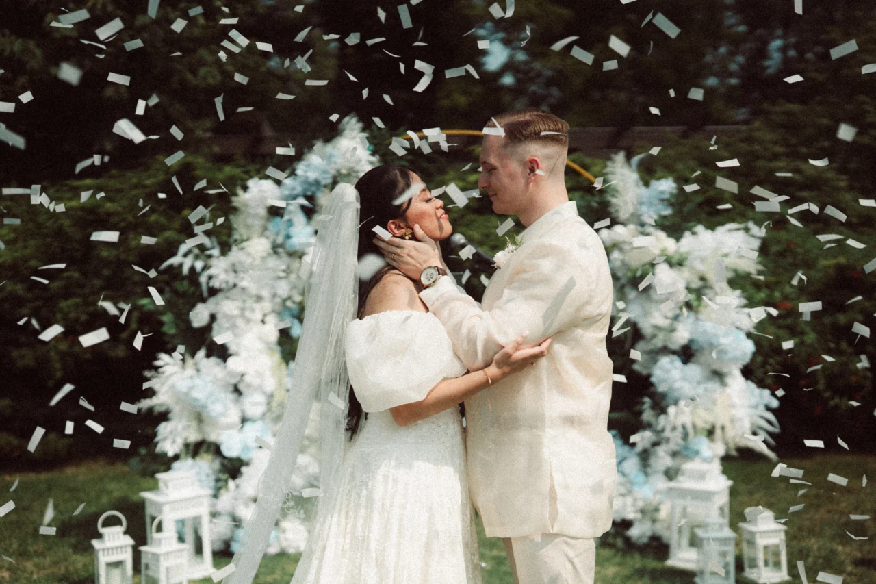 A wedding couple standing in front of an arch of flowers wearing white while confetti flies