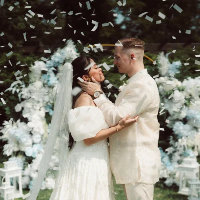 A wedding couple standing in front of an arch of flowers wearing white while confetti flies